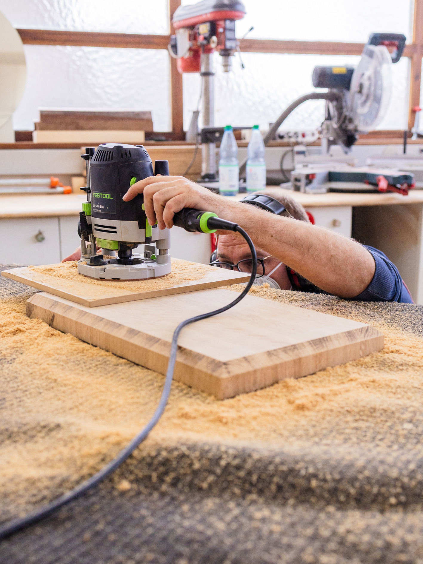 Hände führen eine Oberfräse über eine Holzplatte in der Werkstatt.