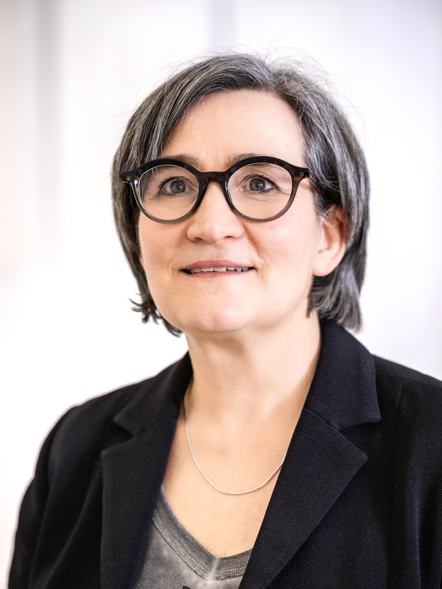 Nahaufnahme von Imke Valentien mit schwarzer Brille und Blazer vor einer hellen Wand.