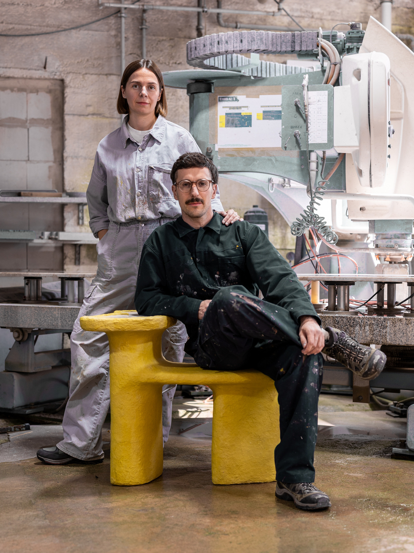 Inna und Marius in Arbeitskleidung mit einem gelben Sitzmöbel vor einer Maschine in der Werkstatt.