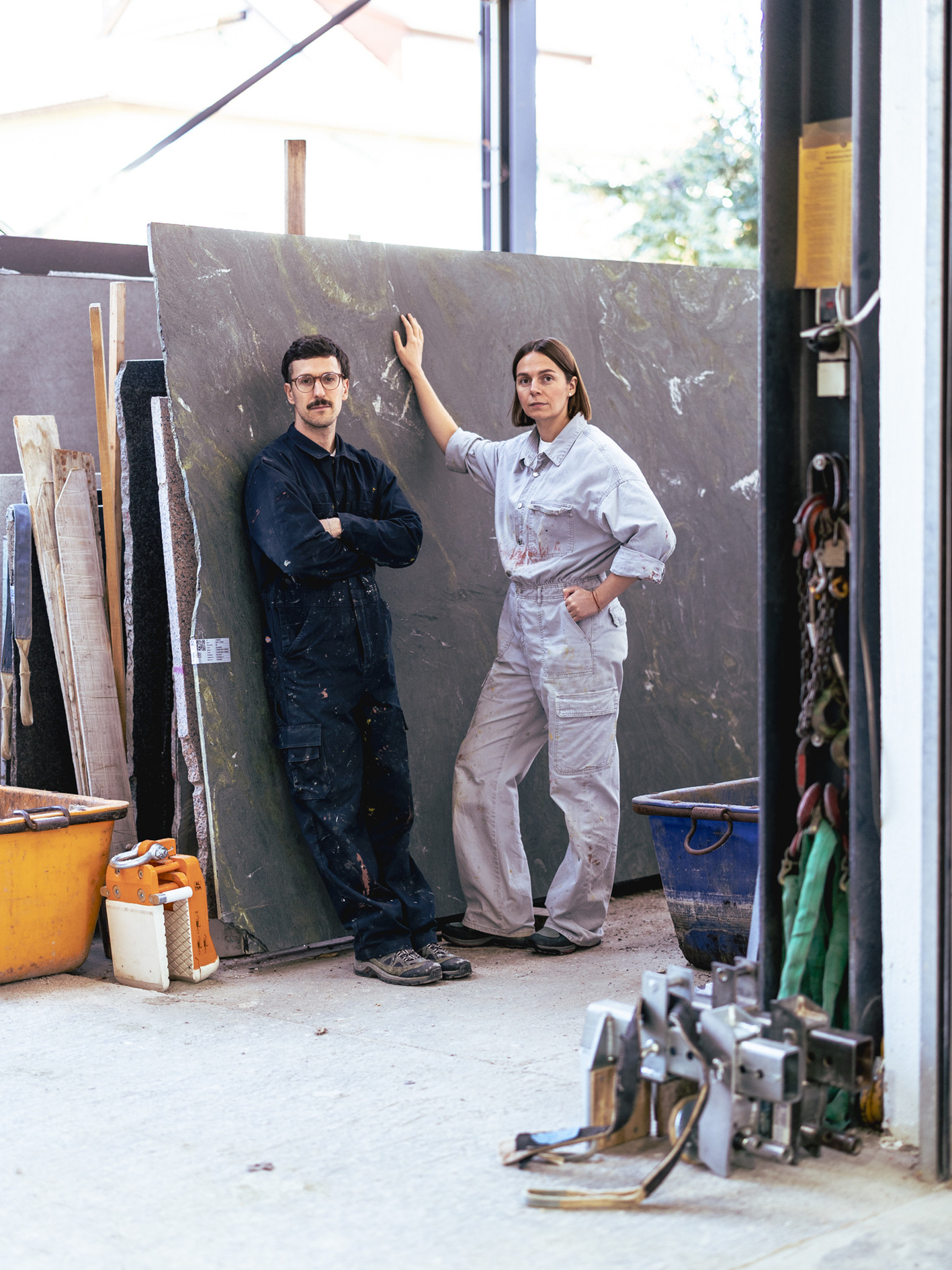 Inna und Marius in Arbeitskleidung vor einer großen Steinplatte in der Werkstatt.